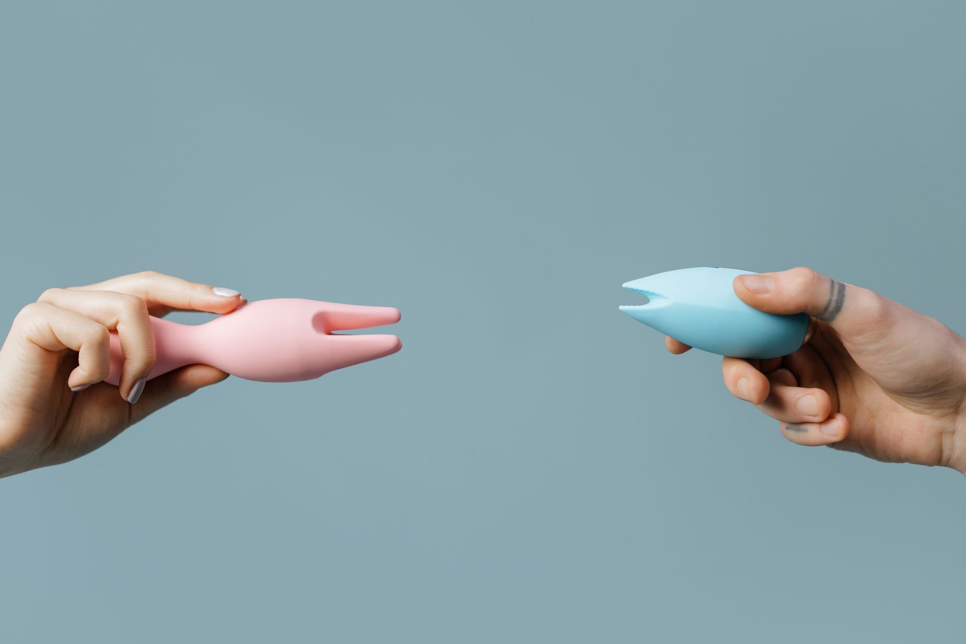 Close-up of two hands holding pink and blue silicone vibrators against a pastel background